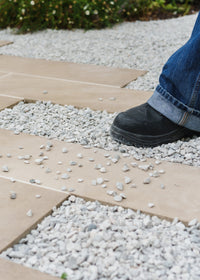 Person wearing blue jeans and black shoes stepping onto a stone patio from gravel